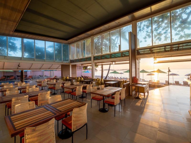 Světla restaurace s dřevěným nábytkem a velkými okny, západ slunce viditelný z terasy.