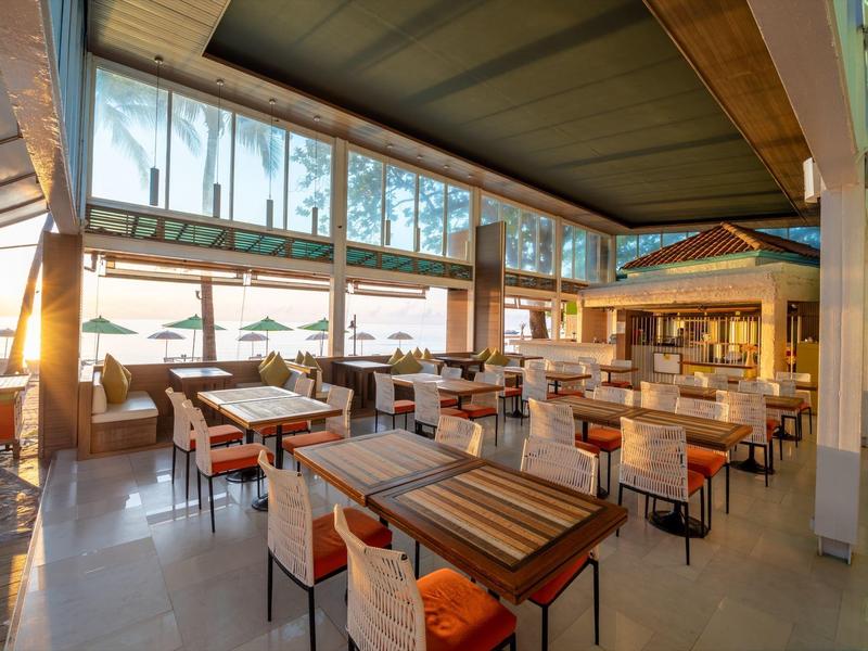 Světelná restaurace s dřevěným nábytkem a velkými okny s výhledem na palmy.