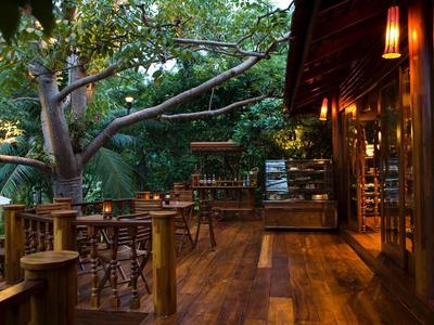 Holzterrasse im Garten mit Tischen und Stühlen, umgeben von Bäumen und sanfter Beleuchtung.