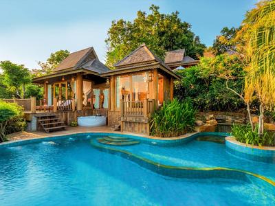 Holzbungalow mit Terrassen und Whirlpool neben einem großen blauen Pool, umgeben von Grün.