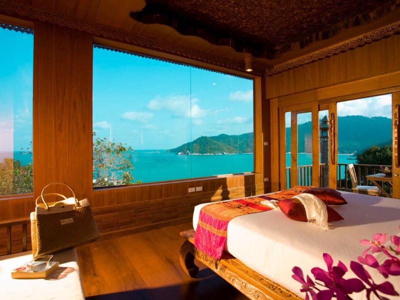 Helles Zimmer mit Holzmöbeln, großen Fenstern und Blick auf blauen See und grüne Berge.