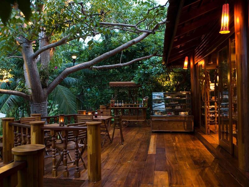 Holzterrasse im Garten mit Tischen und Stühlen, umgeben von Bäumen und sanfter Beleuchtung.