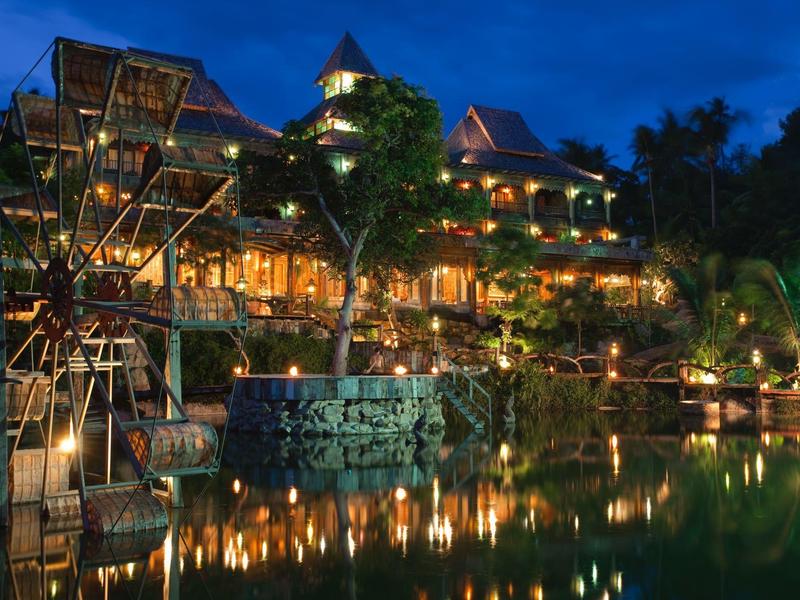 Beleuchtetes Resort mit Wasser, Brücke und Palmen bei Nacht.