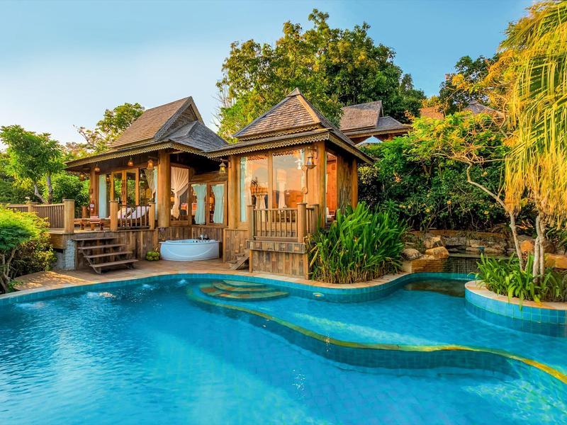 Holzhütte mit einer Terrasse, eingebettet in üppigem Grün, neben einem blauen, geschwungenen Pool.