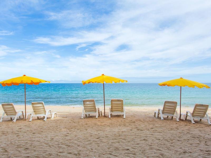 Strand mit sechs Liegestühlen und drei gelben Sonnenschirmen vor blauem Meer und wolkigem Himmel.