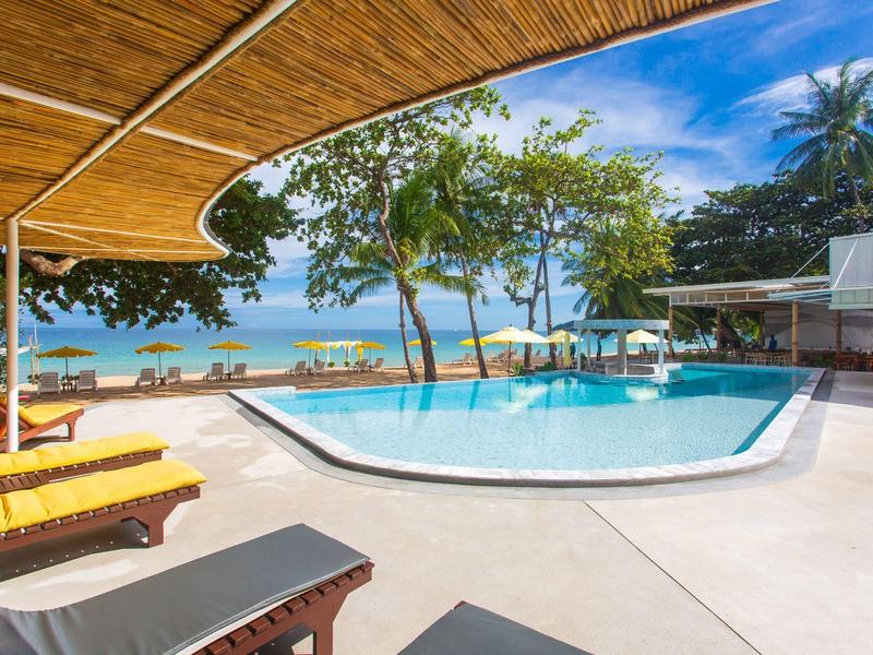 Schwimmbad mit Sonnenschirmen und Liegestühlen, Palmen und Meer im Hintergrund unter blauem Himmel.