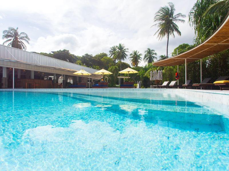 Großer Pool mit klarem Wasser, gelben Sonnenschirmen, Palmen und überdachtem Bereich im Hintergrund.