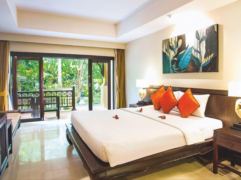 Helles Schlafzimmer mit großem Bett, roten Kissen, Holzmöbeln und Blick auf grünen Balkon.