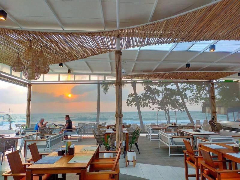 Offenes Restaurant mit Holzdach und Blick auf Meer bei Sonnenuntergang, Tische mit Stühlen und Pflanzen.