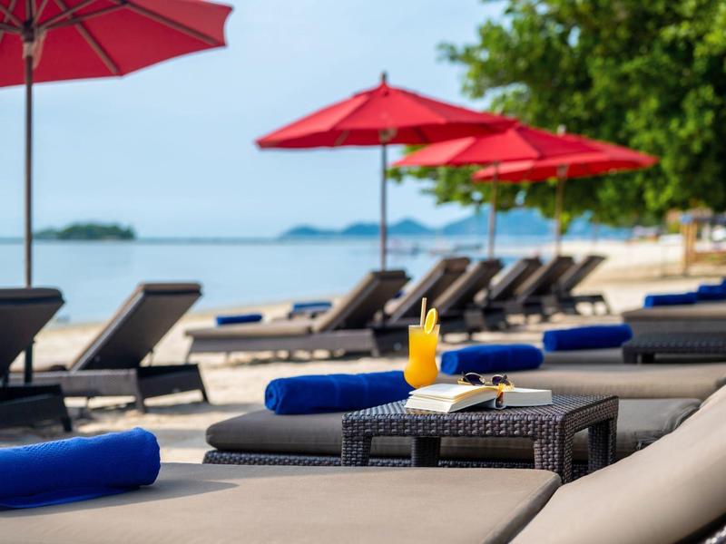 Strand mit Liegen, roten Sonnenschirmen und Getränken bei klarem Himmel und blauem Meer.
