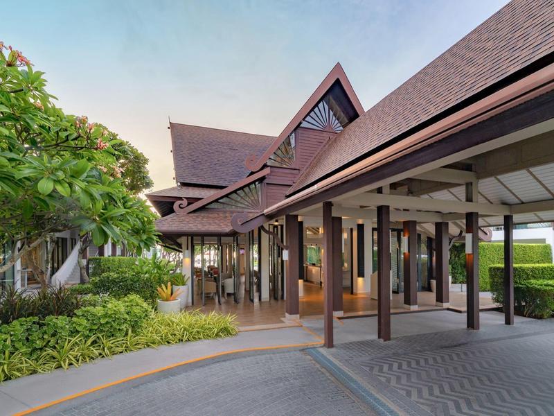 Haus mit spitzem Dach, großen Fenstern und einer überdachten Einfahrt, umgeben von grünen Pflanzen.