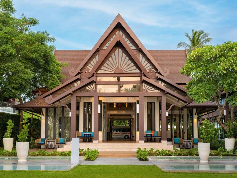 Traditionelles thailändisches Holzhaus mit spitzem Dach, umgeben von grünen Bäumen und klarem Wasser.