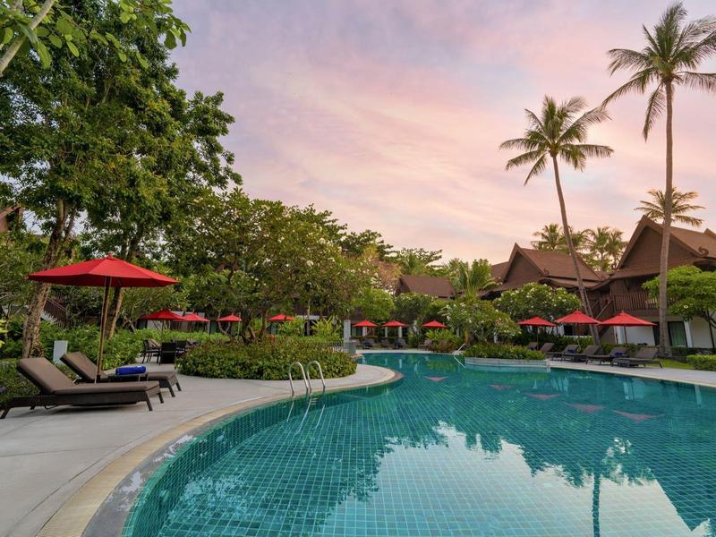 Runder Pool umgeben von Liegestühlen, roten Sonnenschirmen und Palmen bei Sonnenuntergang.