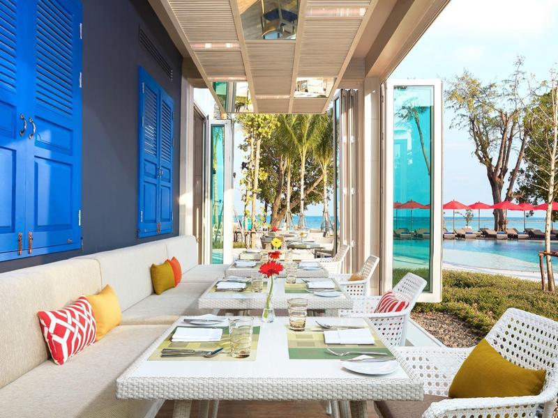 Moderner Außenbereich eines Restaurants mit weißen Tischen, Stühlen und blauen Fensterläden am Wasser.