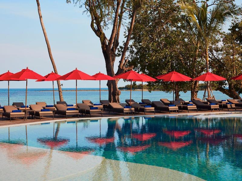 Einladender Pool mit roten Sonnenschirmen und Liegestühlen am Ufer, umgeben von Bäumen.