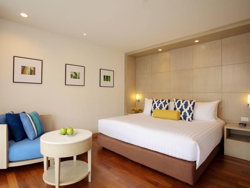 Hell eingerichtetes Hotelzimmer mit Doppelbett, beigem Sofa, rundem Tisch und Wandbildern.