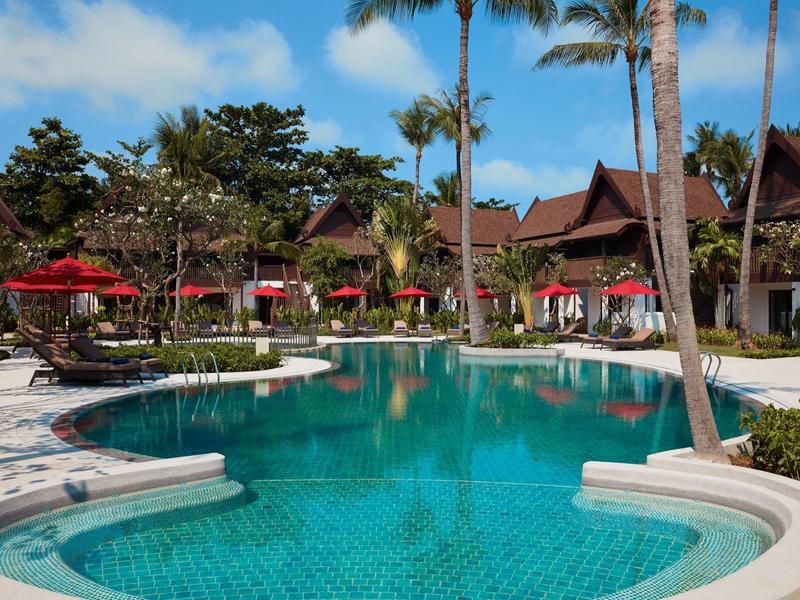 Ein tropischer Außenpool mit Palmen, roten Sonnenschirmen und traditionellen Holzhäusern im Hintergrund.