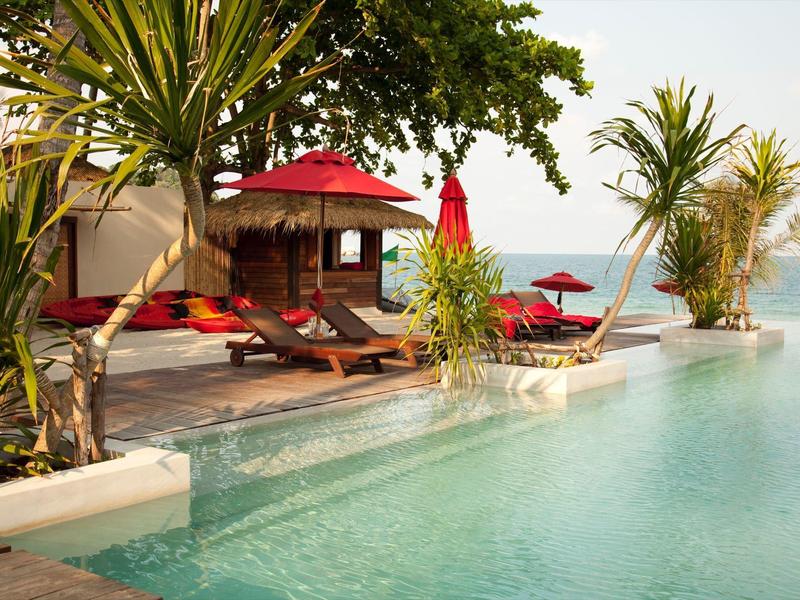 Einladender Pool mit rotem Sonnenschirm, zwei Liegen und Palmen am Rand bei klarem Himmel.