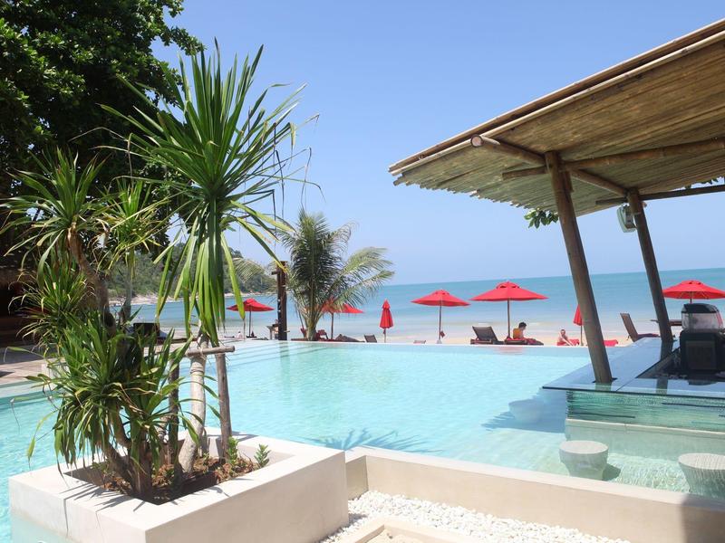 Ein Swimmingpool mit grünen Pflanzen, roten Sonnenschirmen und Strand im Hintergrund.