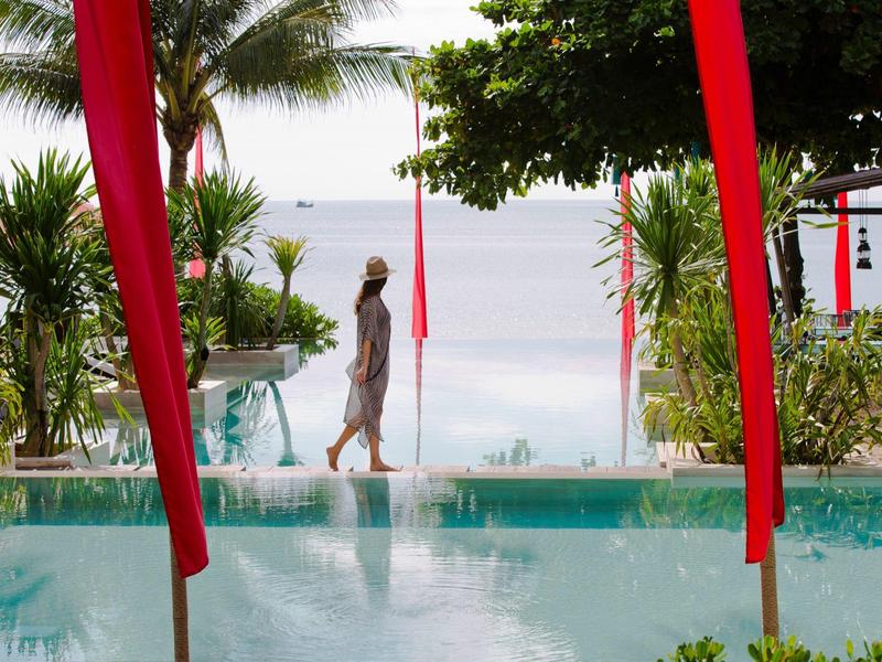 Frau geht am Pool entlang, umgeben von grünen Palmen und roten Vorhängen, Blick aufs Meer.