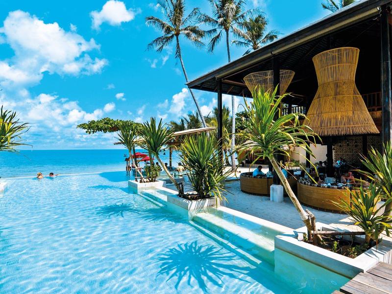 Infinity-Pool am Meer mit Palmen, blauer Himmel und modernem, offenem Lounge-Bereich.