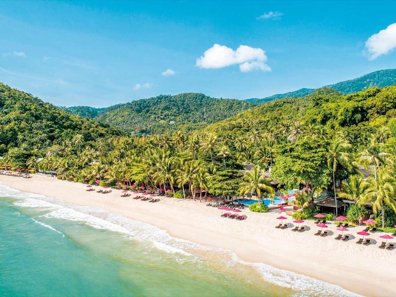 Ein breiter Sandstrand mit grünen Hügeln, Palmen und blauem Himmel mit wenigen Wolken.