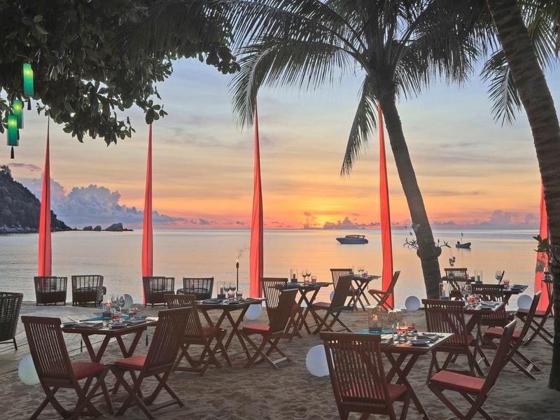 Strandrestaurant mit Tischen, Stühlen und Palmen bei Sonnenuntergang am Meer.