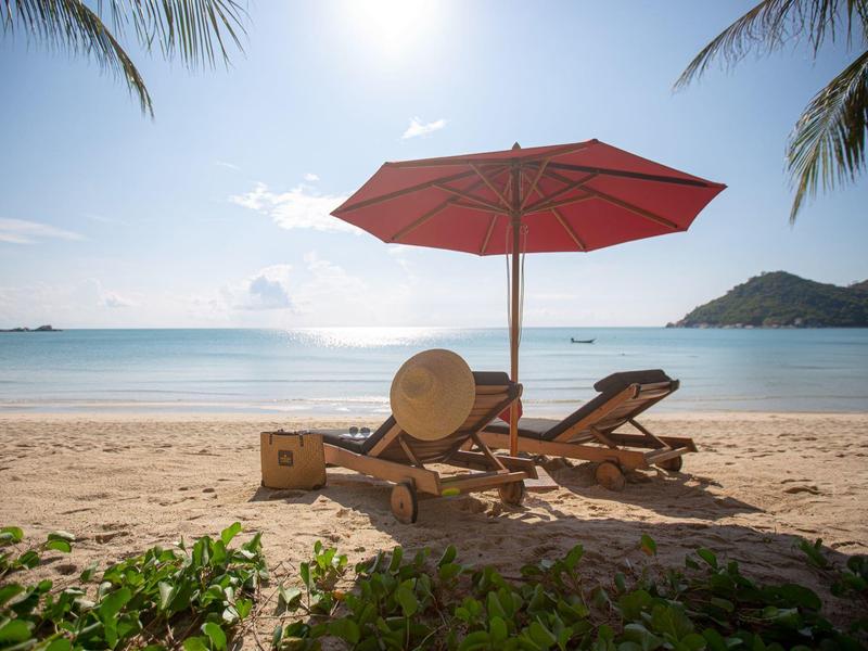 Strand mit zwei Holzliegen unter rotem Sonnenschirm, Palmen, ruhiges Meer und heller Himmel.