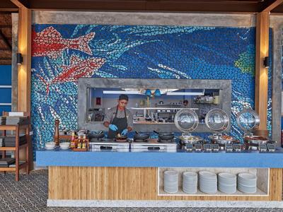 Buffet mit blauer Mosaikwand, Meeresmotiv, verschiedenen Speisen und Schüsseln auf der Theke.