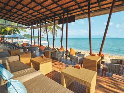 Offene Strandbar mit Holzdach, Sitzgruppen aus Holz und Korbmöbeln, Blick auf Meer und Palmen.