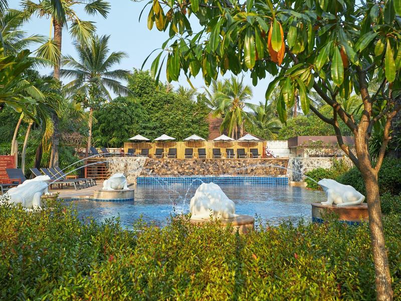 Pool mit Liegestühlen und Sonnenschirmen, umgeben von tropischen Pflanzen und Bäumen