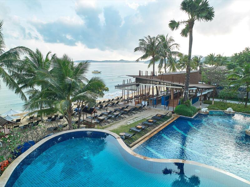 Blick auf einen tropischen Hotelpool mit Palmen und Meer im Hintergrund.