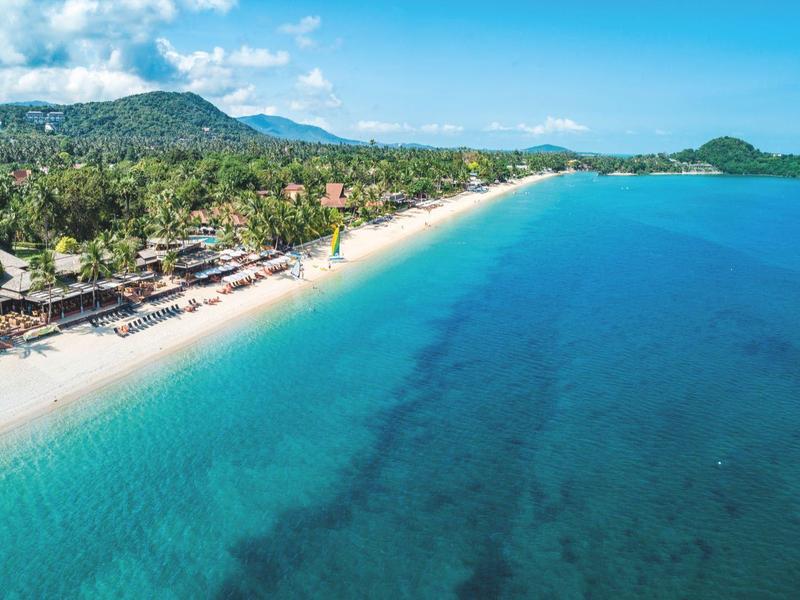Langer Sandstrand mit klarem, blauem Wasser und tropischer Vegetation im Hintergrund