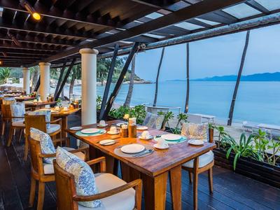 Restaurant mit Holztischen und weißen Sitzpolstern, großer Glasfront mit Blick aufs Meer und Berge.