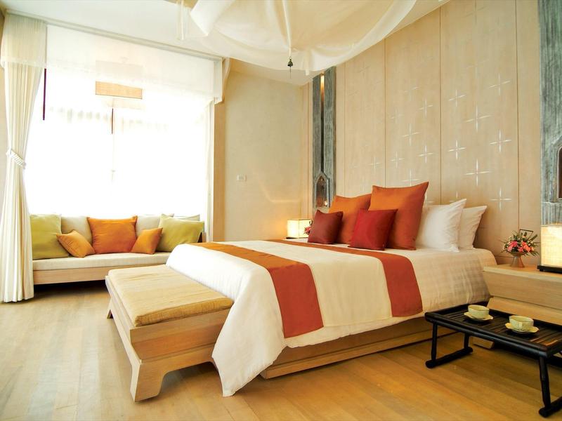 Světý hotelový pokoj s velkou postelí, barevnými polštáři a útulným posezením u okna.