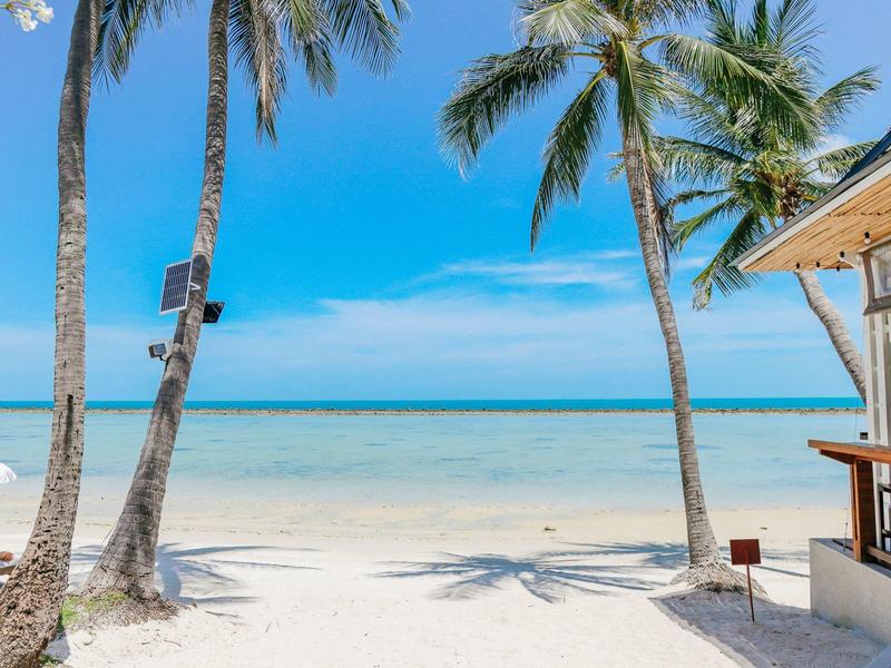 Strand mit weißem Sand, Palmen und einer kleinen Bar am Meer unter blauem Himmel.