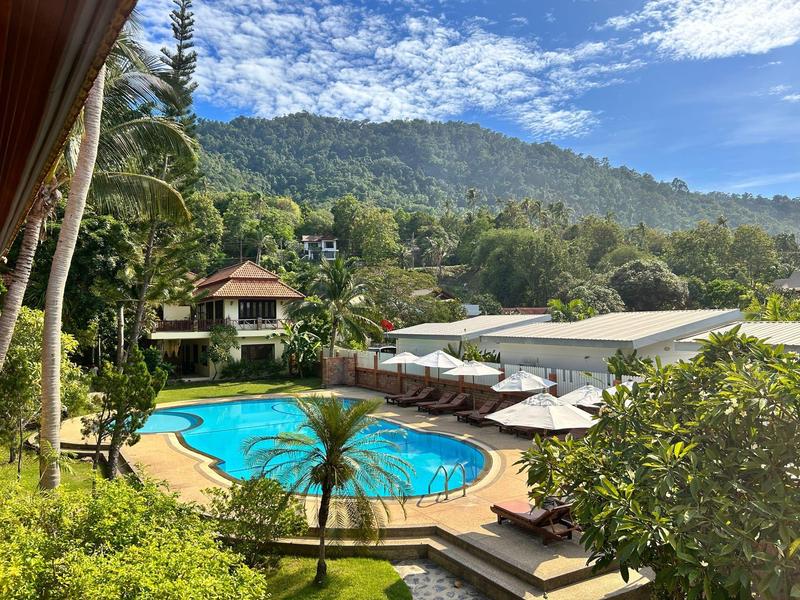 Rundes Schwimmbecken mit Sonnenschirmen, tropische Pflanzen und bewaldeter Hügel im Hintergrund.