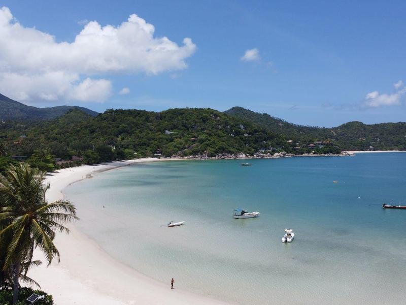 Strand mit weißem Sand, türkisfarbenem Wasser, einigen Booten und bewaldeten Hügeln im Hintergrund.