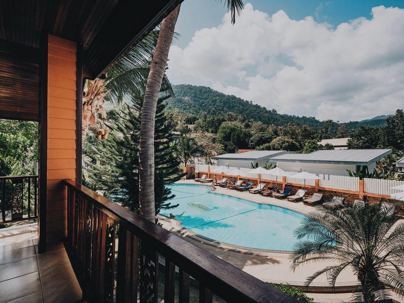 Blick von Balkon auf blauen Pool, Liegestühle, Palmen und grüne Hügel unter blauem Himmel.