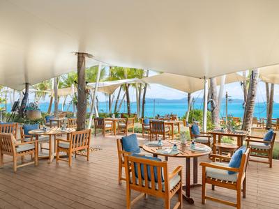 Otwarta jadalnia z drewnianymi stołami i krzesłami, widok na palmy i ocean.