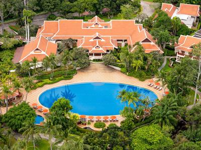 Widok z lotu ptaka na ośrodek z dużym basenem otoczonym tropikalnymi roślinami i budynkami z czerwonymi dachami.