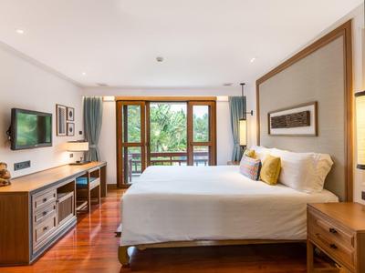 Jasny pokój hotelowy z dużym łóżkiem, telewizorem, biurkiem i balkonem z widokiem na zieleń.