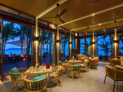 Otwarta restauracja z drewnianymi meblami i widokiem na palmy oraz ocean o zachodzie słońca.