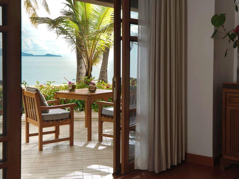 Widok przez drzwi na taras z stołem i krzesłami, palmami i oceanem w tle.