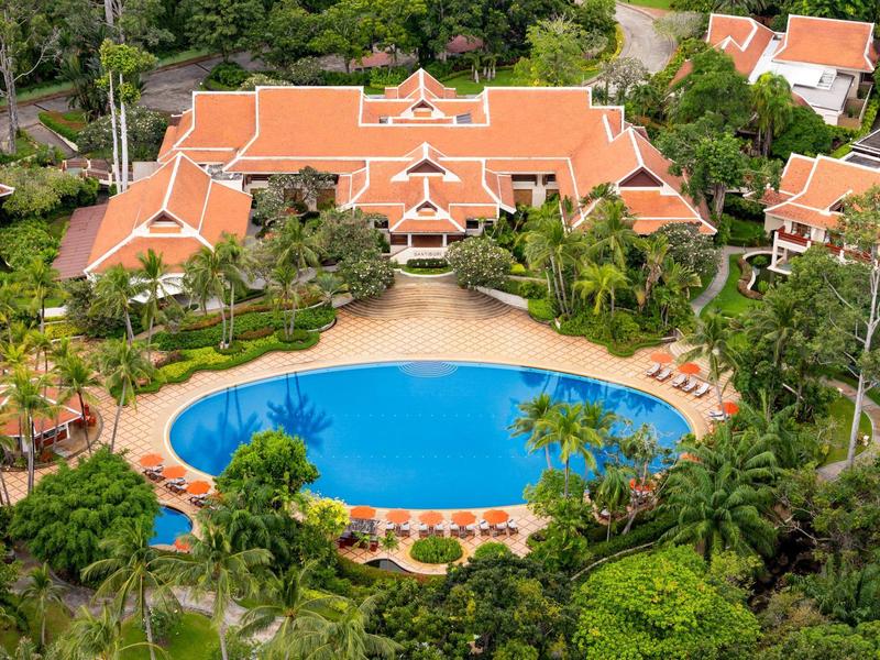 Ośrodek hotelowy z dużym, owalnym basenem otoczonym leżakami i tropikalną roślinnością.