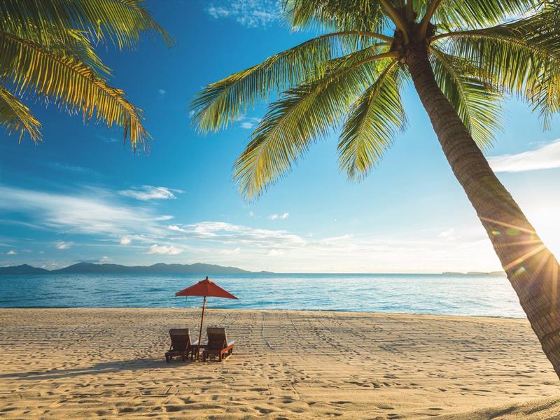 Plaża z palmami, parasolem i dwoma krzesłami przy spokojnym morzu o zachodzie słońca.