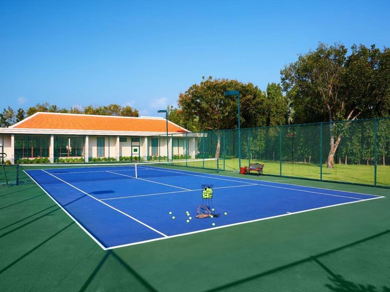 Czysty kort tenisowy z niebieską nawierzchnią i małym budynkiem w tle w słoneczny dzień.