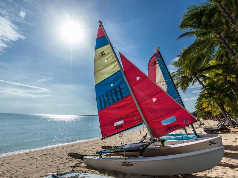 Żaglówki z kolorowymi żaglami stoją na tropikalnej piaszczystej plaży pod palmami.
