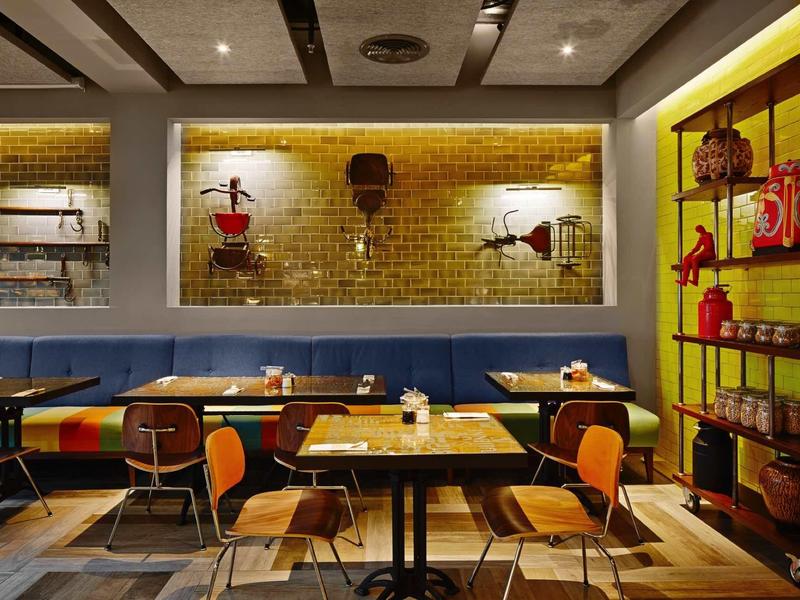 Modern restaurant with wooden tables, colorful chairs, and wall decorations with warm lighting.