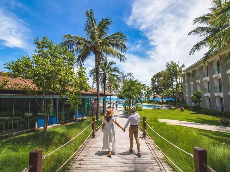 Couple holding hands on wooden boardwalk between tropical plants leading to a seaside resort.